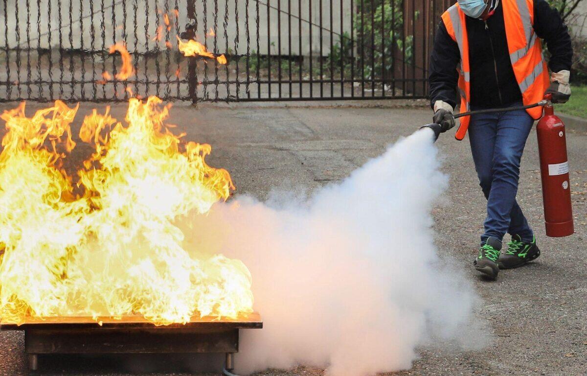Person in a safety vest uses a fire extinguisher to put out a large blaze on a metal tray outdoors, with thick white smoke nearby.