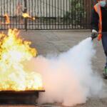 Person in a safety vest uses a fire extinguisher to put out a large blaze on a metal tray outdoors, with thick white smoke nearby.