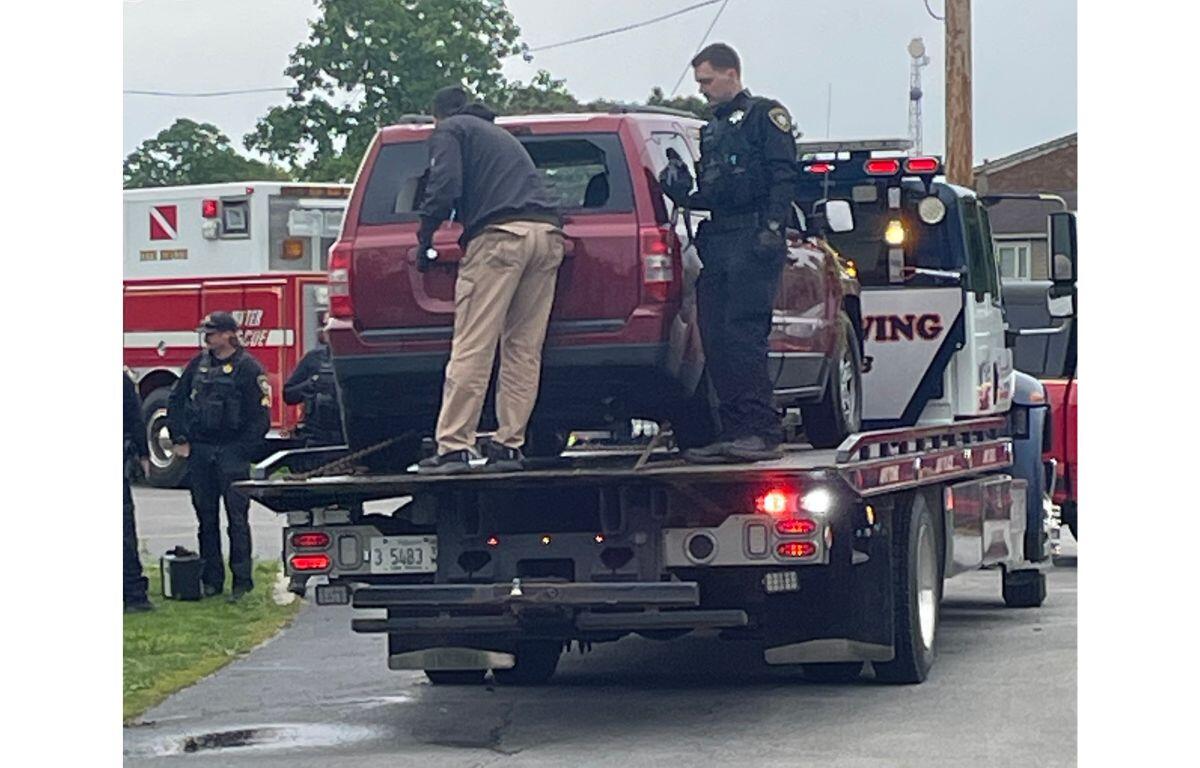 Tow truck carrying a red SUV with a uniformed officer and another responder on scene on a residential street, ambulance in the background.