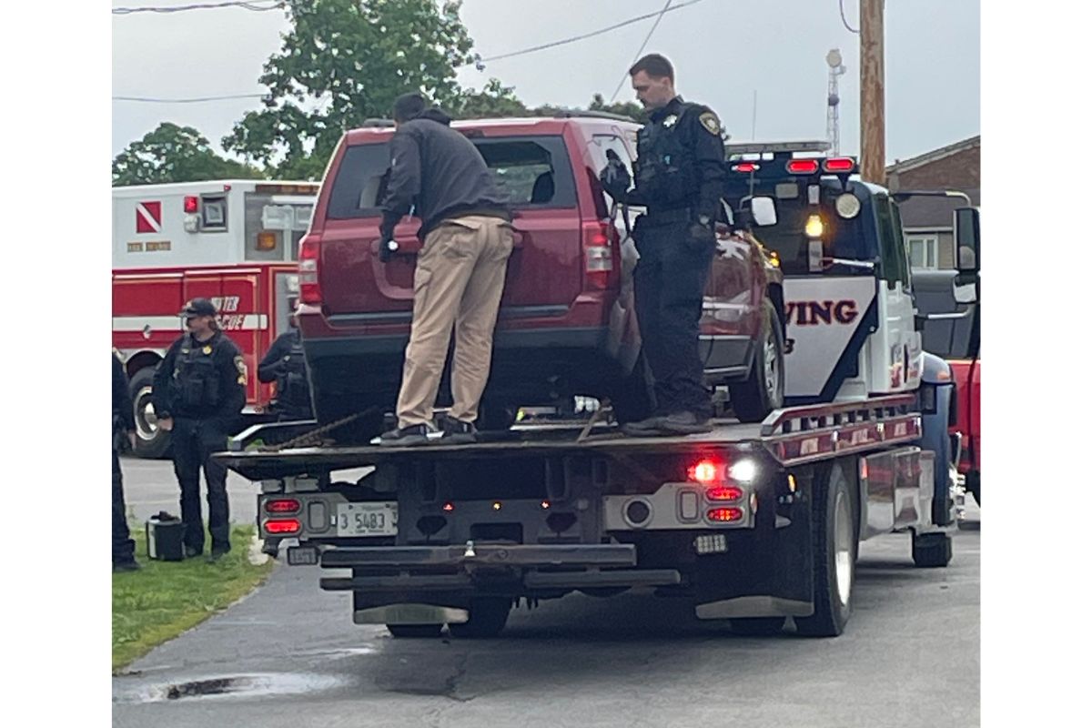 Tow truck carrying a red SUV with a uniformed officer and another responder on scene on a residential street, ambulance in the background.