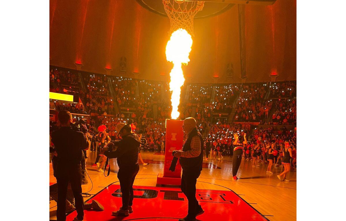 Basketball arena crowd watches a large flame erupting from a device at center court during a halftime show.