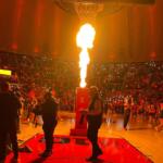 Basketball arena crowd watches a large flame erupting from a device at center court during a halftime show.