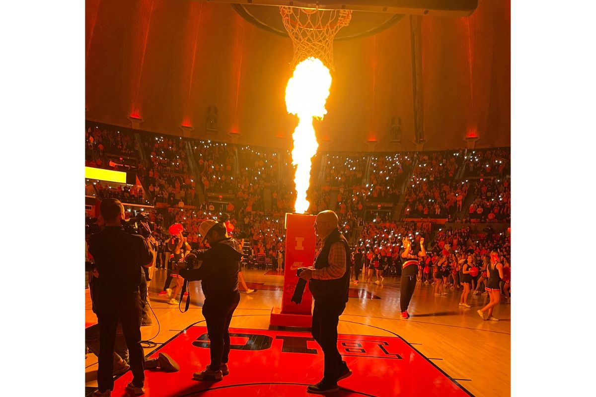 Basketball arena crowd watches a large flame erupting from a device at center court during a halftime show.