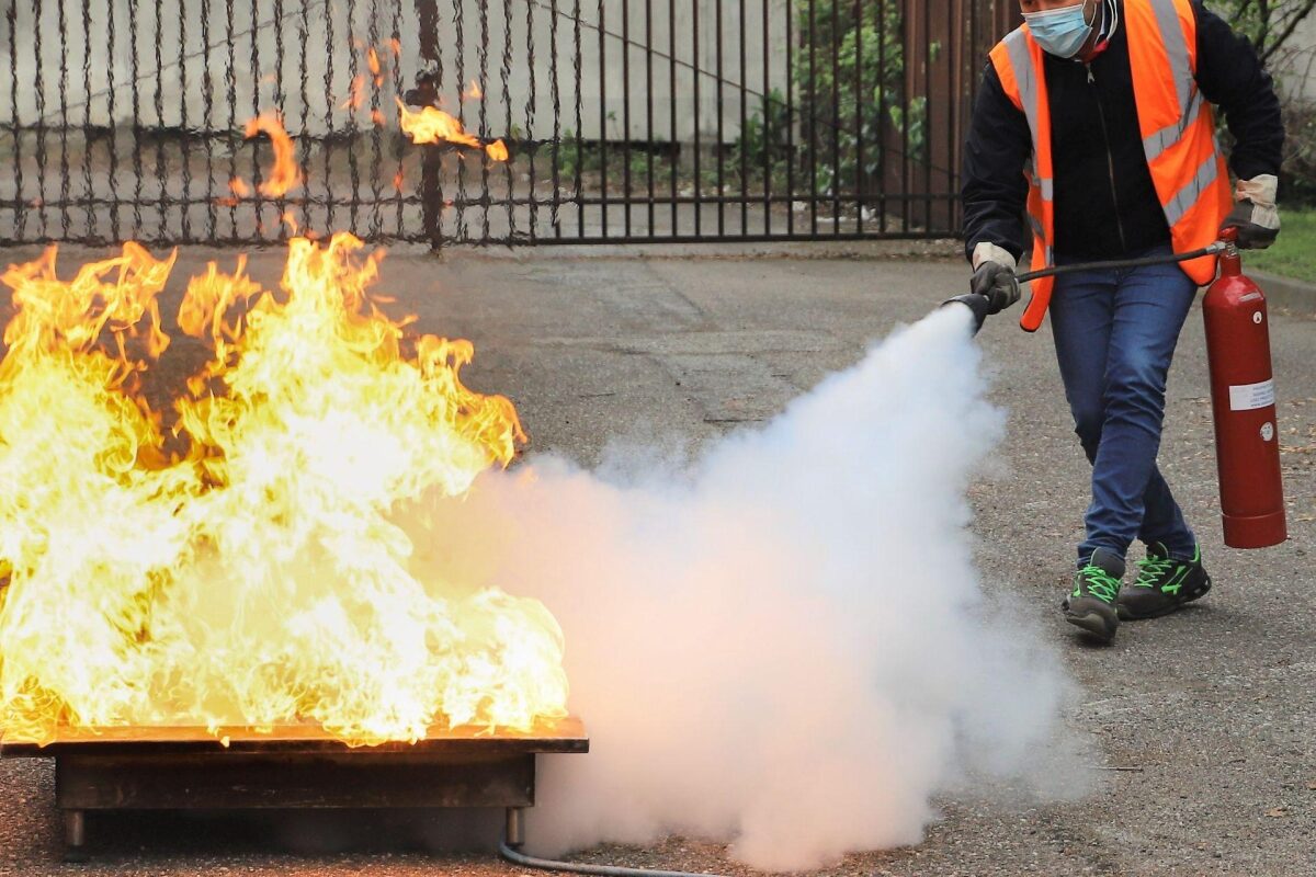 Person in a safety vest uses a fire extinguisher to put out a large blaze on a metal tray outdoors, with thick white smoke nearby.