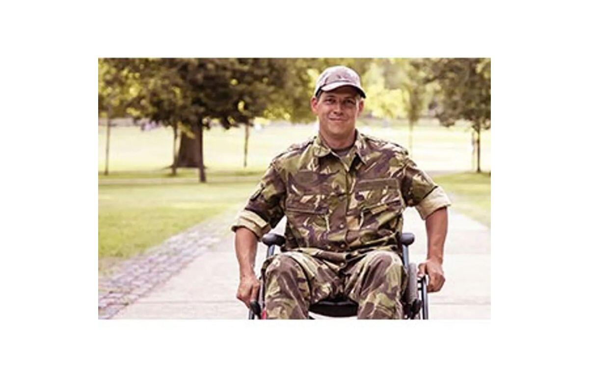 Man in camouflage military uniform in a wheelchair on a park path, smiling at the camera