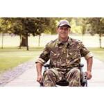 Man in camouflage military uniform in a wheelchair on a park path, smiling at the camera