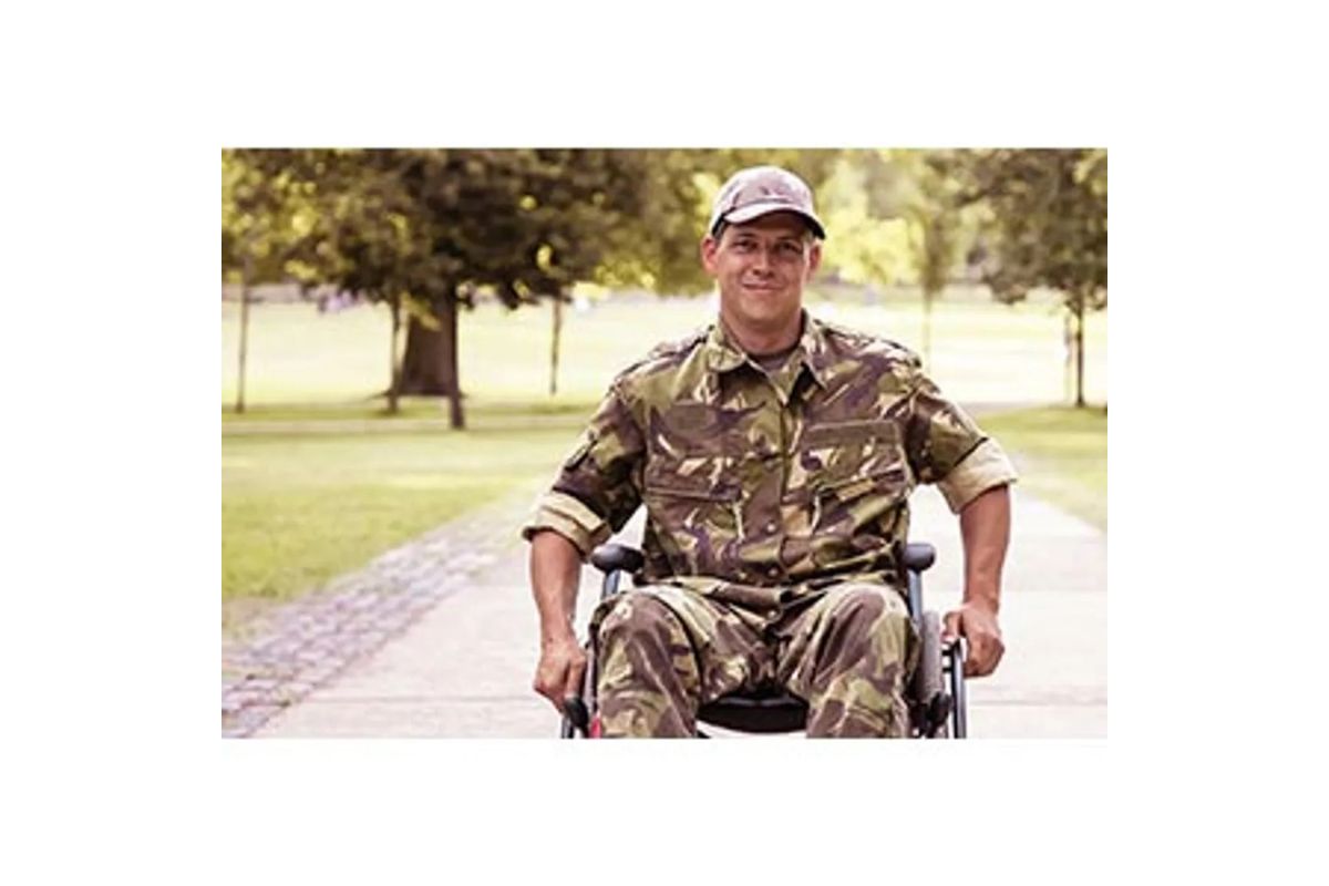 Man in camouflage military uniform in a wheelchair on a park path, smiling at the camera