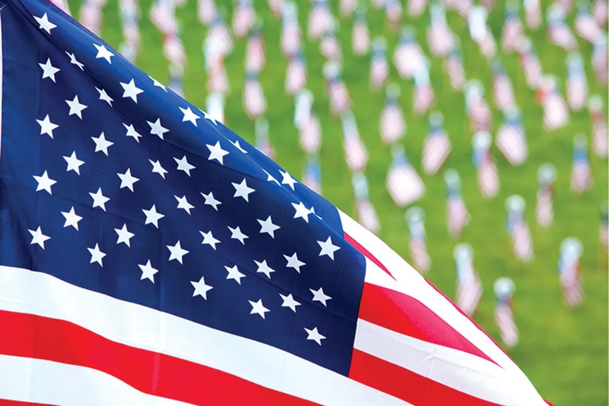 Close-up of a waving United States flag in the foreground, with many small American flags planted on a green lawn in the blurred background.