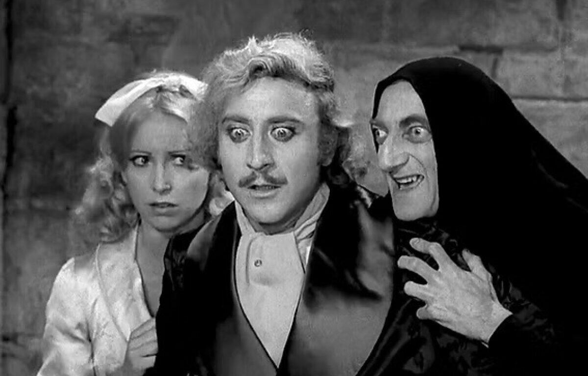 Three actors in a black-and-white scene: a startled man in the center, a worried woman on the left, and a hooded sinister figure on the right.