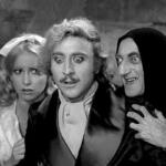 Three actors in a black-and-white scene: a startled man in the center, a worried woman on the left, and a hooded sinister figure on the right.