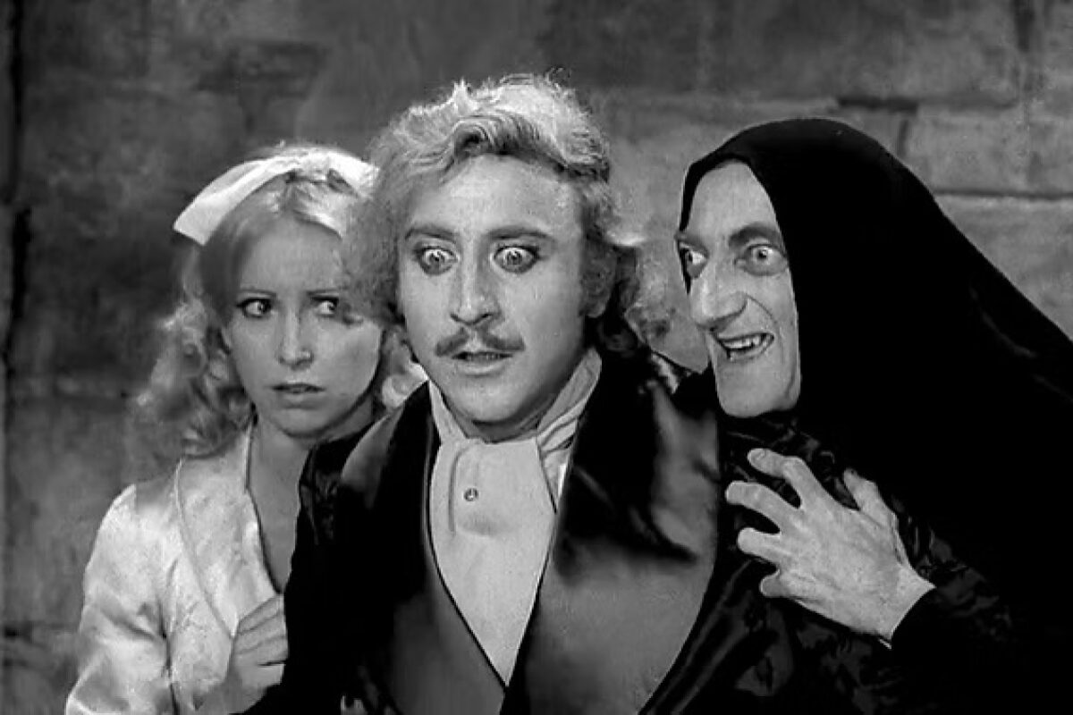 Three actors in a black-and-white scene: a startled man in the center, a worried woman on the left, and a hooded sinister figure on the right.