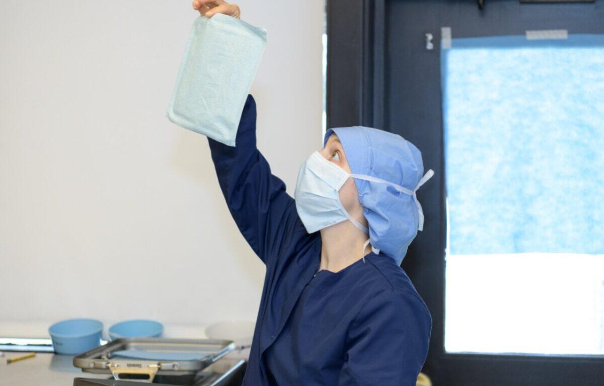Medical professional in scrubs and a mask holds a sterile blue pad up toward the ceiling in a clinical room.
