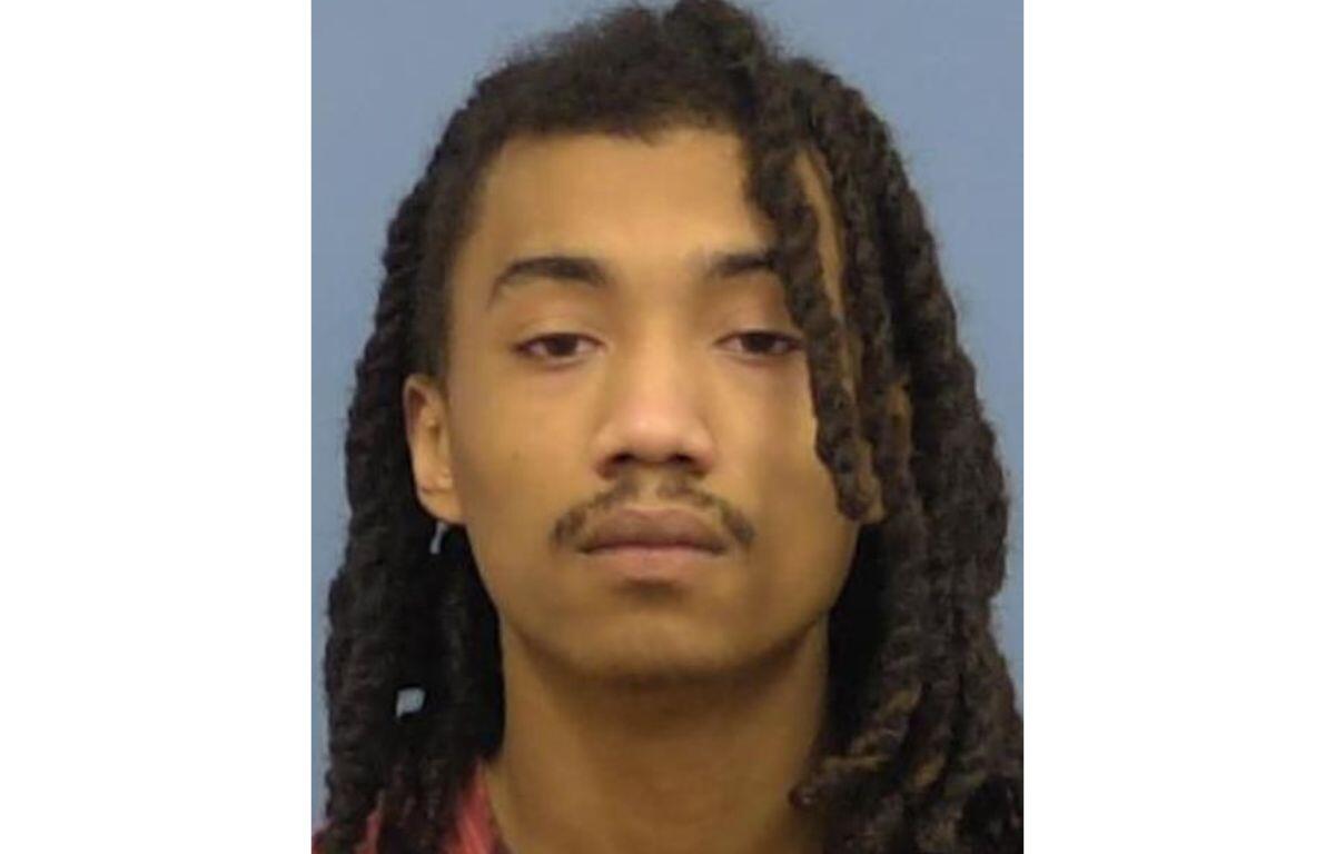 Mugshot-style portrait of a young man with long dreadlocks, neutral expression, wearing a red shirt against a blue background.