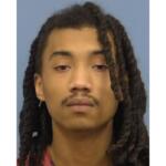 Mugshot-style portrait of a young man with long dreadlocks, neutral expression, wearing a red shirt against a blue background.