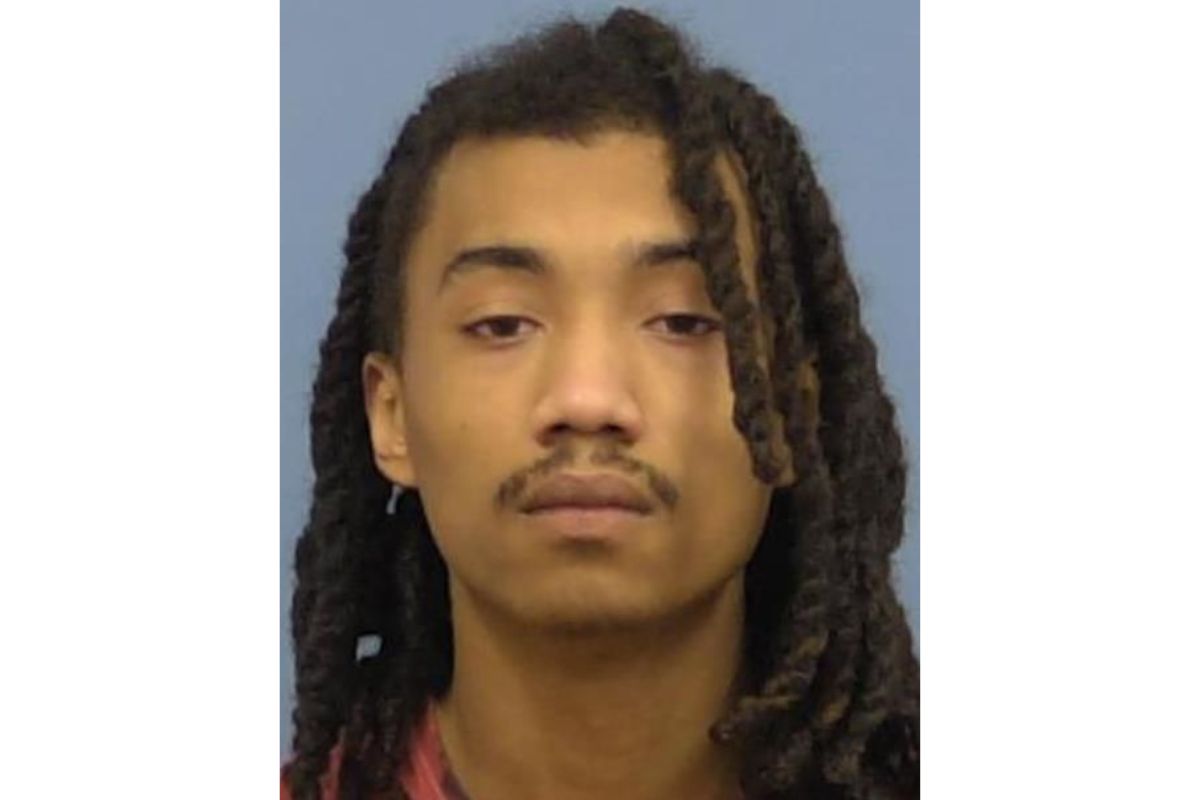 Mugshot-style portrait of a young man with long dreadlocks, neutral expression, wearing a red shirt against a blue background.