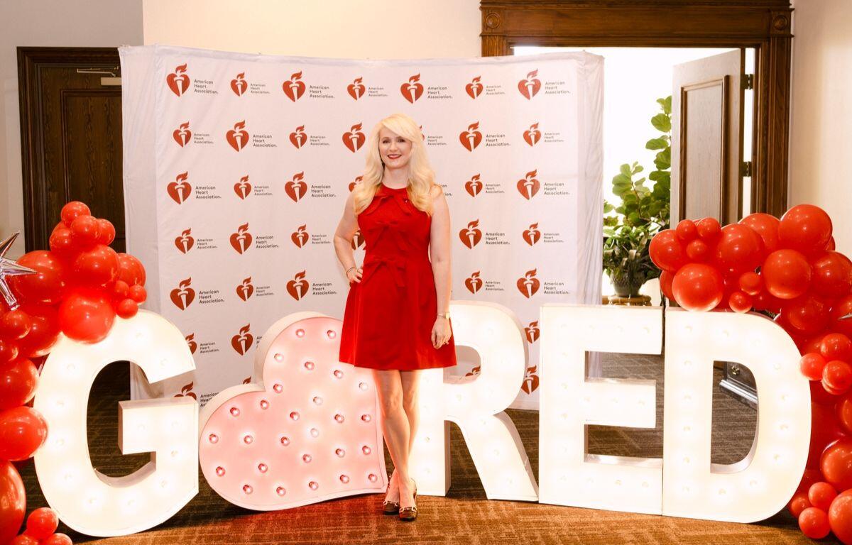 Woman in a red dress posing among oversized illuminated letters and red balloon clusters at an American Heart Association event backdrop.