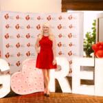 Woman in a red dress posing among oversized illuminated letters and red balloon clusters at an American Heart Association event backdrop.