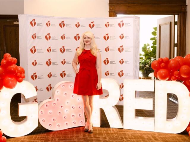 Woman in a red dress posing among oversized illuminated letters and red balloon clusters at an American Heart Association event backdrop.