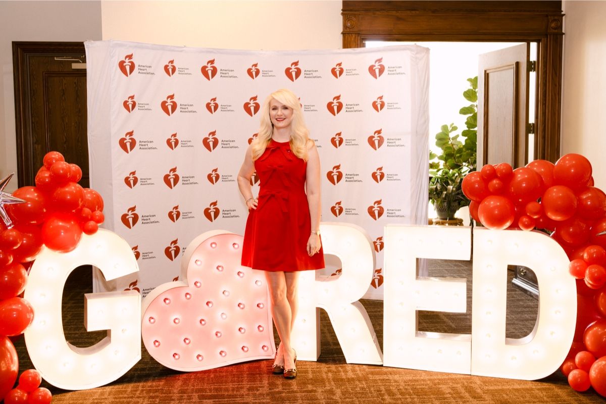 Woman in a red dress posing among oversized illuminated letters and red balloon clusters at an American Heart Association event backdrop.