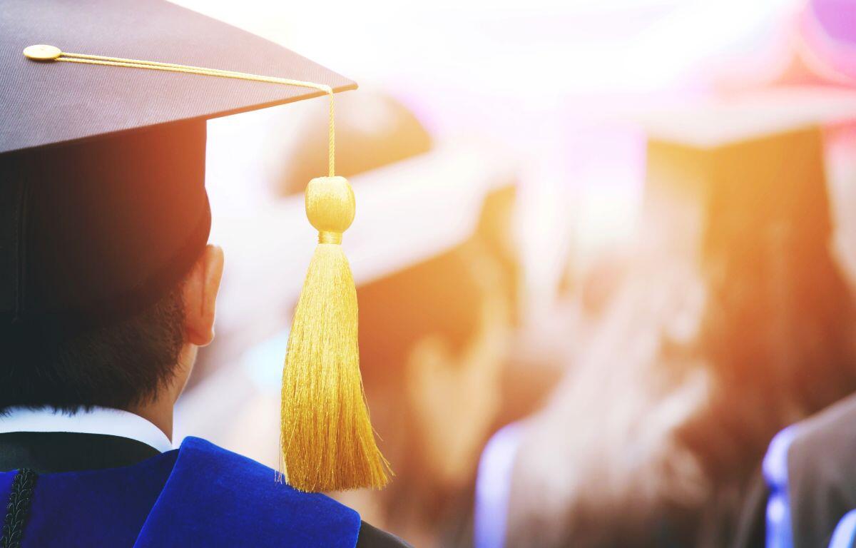 Back view of a graduate in a cap and blue gown, tassel hanging from the cap at a commencement ceremony