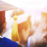 Back view of a graduate in a cap and blue gown, tassel hanging from the cap at a commencement ceremony