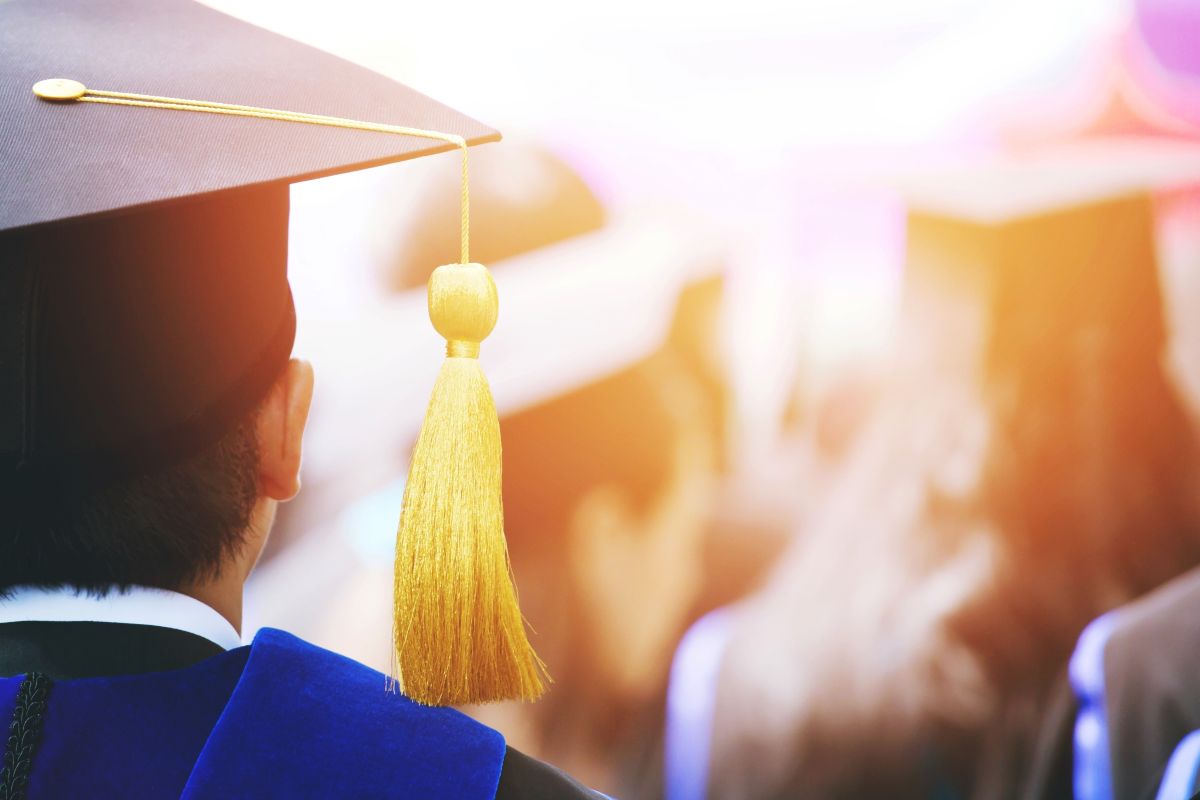 Back view of a graduate in a cap and blue gown, tassel hanging from the cap at a commencement ceremony