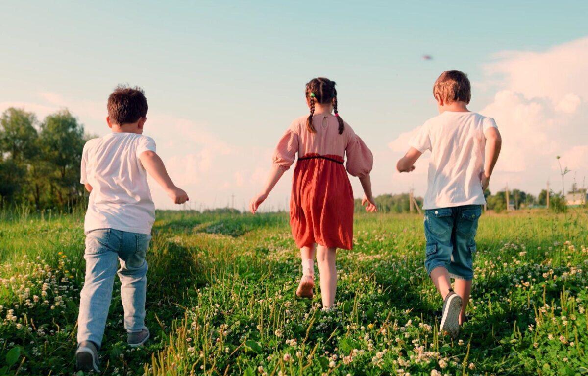 Three children run through a sunny, grassy field with daisies, seen from behind.