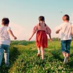 Three children run through a sunny, grassy field with daisies, seen from behind.