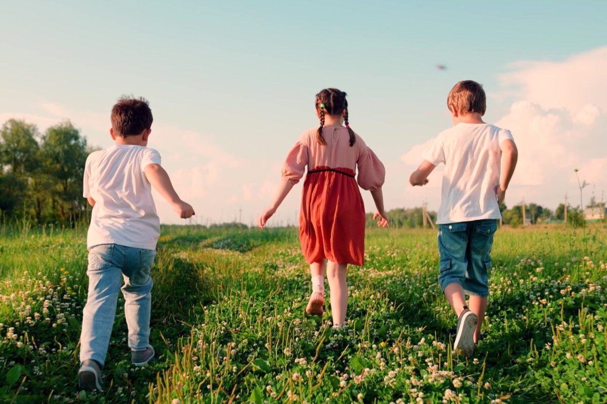 Three children run through a sunny, grassy field with daisies, seen from behind.