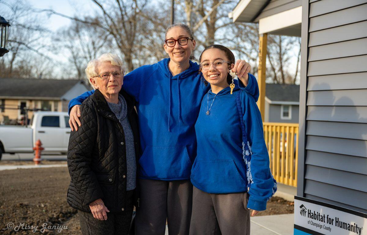 Three women stand together outside a house—an elderly woman in a black quilted jacket flanks two younger women in blue hoodies, smiling as one holds up a key; Habitat for Humanity sign visible nearby.