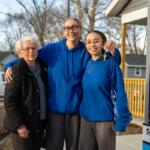 Three women stand together outside a house—an elderly woman in a black quilted jacket flanks two younger women in blue hoodies, smiling as one holds up a key; Habitat for Humanity sign visible nearby.