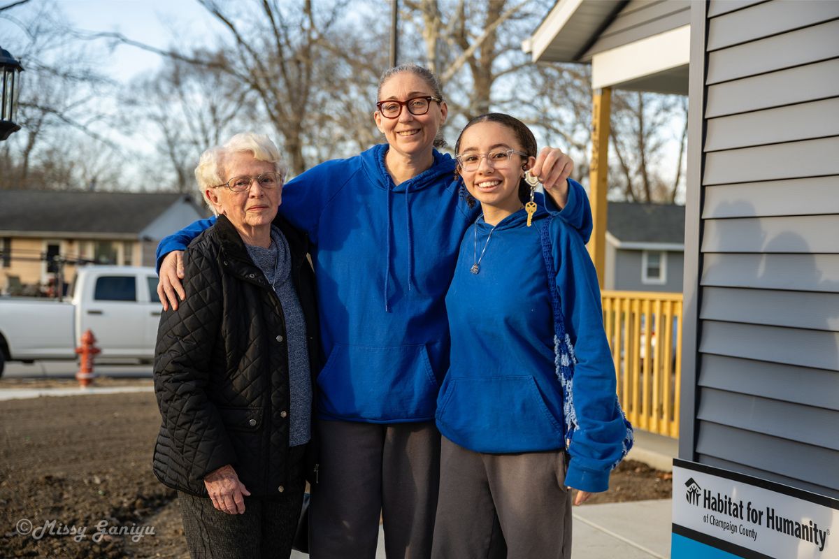 Three women stand together outside a house—an elderly woman in a black quilted jacket flanks two younger women in blue hoodies, smiling as one holds up a key; Habitat for Humanity sign visible nearby.