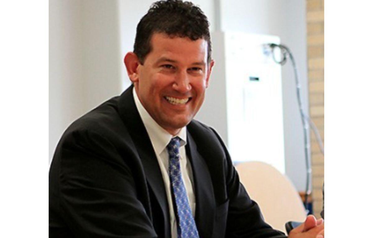 Smiling man in a dark suit and blue patterned tie seated at a table in an office setting.