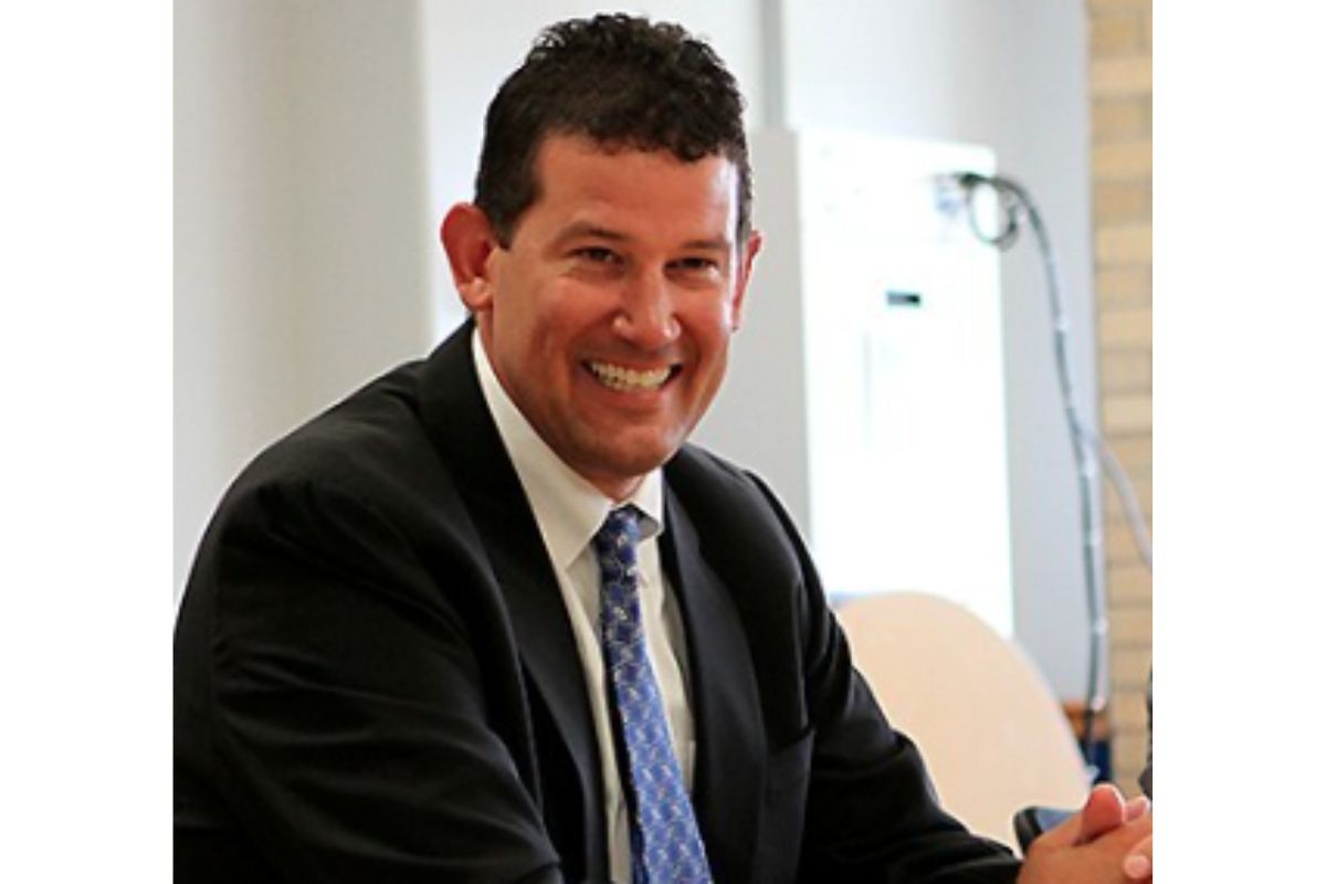 Smiling man in a dark suit and blue patterned tie seated at a table in an office setting.