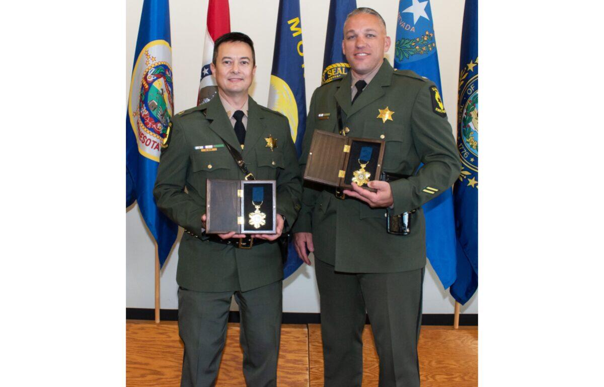 Two military officers in dress greens pose with medal awards in presentation boxes, standing in front of flags backdropped in a ceremony setting.”,