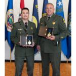 Two military officers in dress greens pose with medal awards in presentation boxes, standing in front of flags backdropped in a ceremony setting.”,