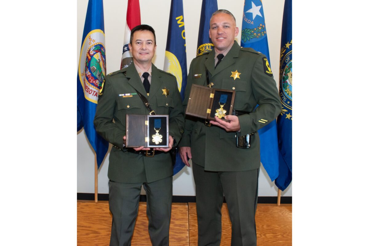 Two military officers in dress greens pose with medal awards in presentation boxes, standing in front of flags backdropped in a ceremony setting.”,