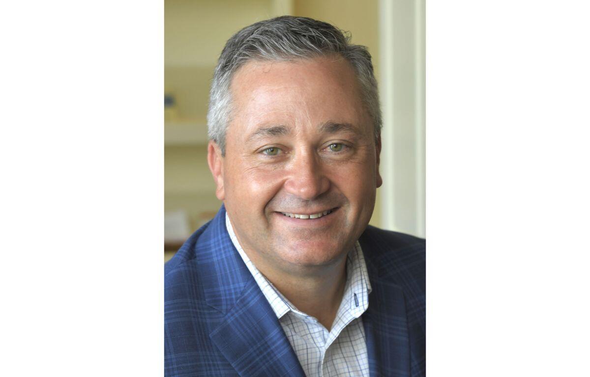 Smiling middle-aged man with short gray hair wearing a blue plaid blazer and white checkered shirt.