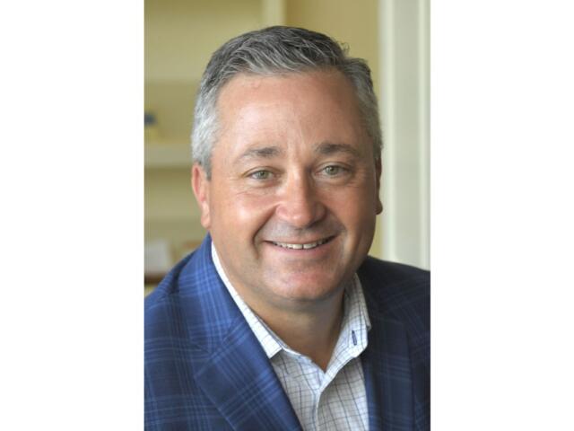 Smiling middle-aged man with short gray hair wearing a blue plaid blazer and white checkered shirt.
