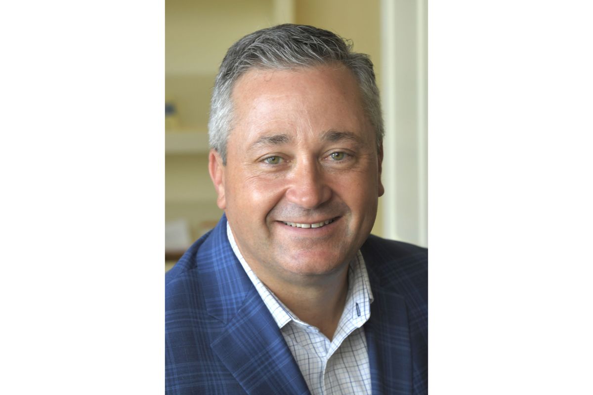 Smiling middle-aged man with short gray hair wearing a blue plaid blazer and white checkered shirt.