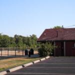 Red wooden concession-style building with a sloped roof beside a paved parking lot under a clear blue sky.
