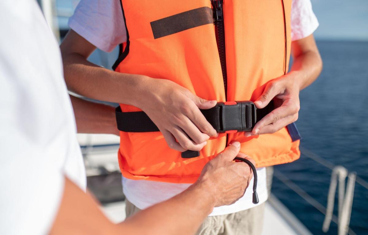 Person fastening a bright orange life jacket on a boat, buckling the strap with the ocean in the background.