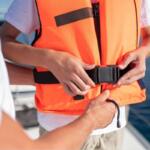 Person fastening a bright orange life jacket on a boat, buckling the strap with the ocean in the background.