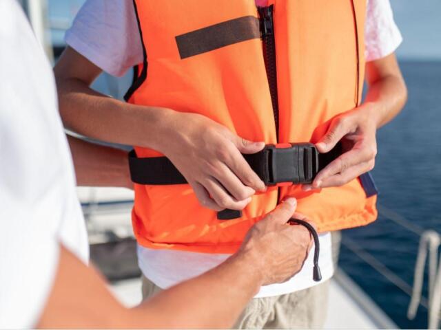 Person fastening a bright orange life jacket on a boat, buckling the strap with the ocean in the background.
