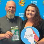 Two smiling adults pose in front of an Illinois Lottery backdrop; man holds a scratch-off ticket, woman holds a round 'I WON!' sign with the Illinois silhouette and colorful stripes.
