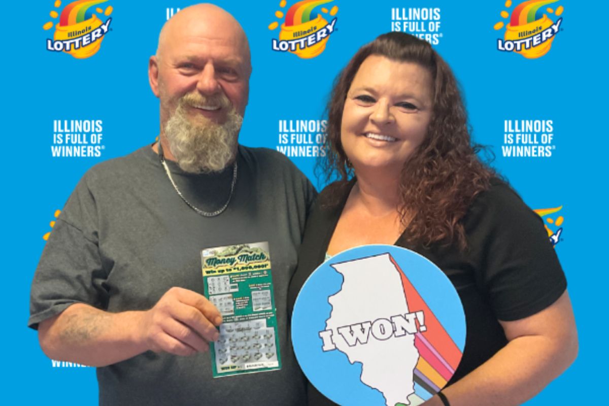 Two smiling adults pose in front of an Illinois Lottery backdrop; man holds a scratch-off ticket, woman holds a round 'I WON!' sign with the Illinois silhouette and colorful stripes.