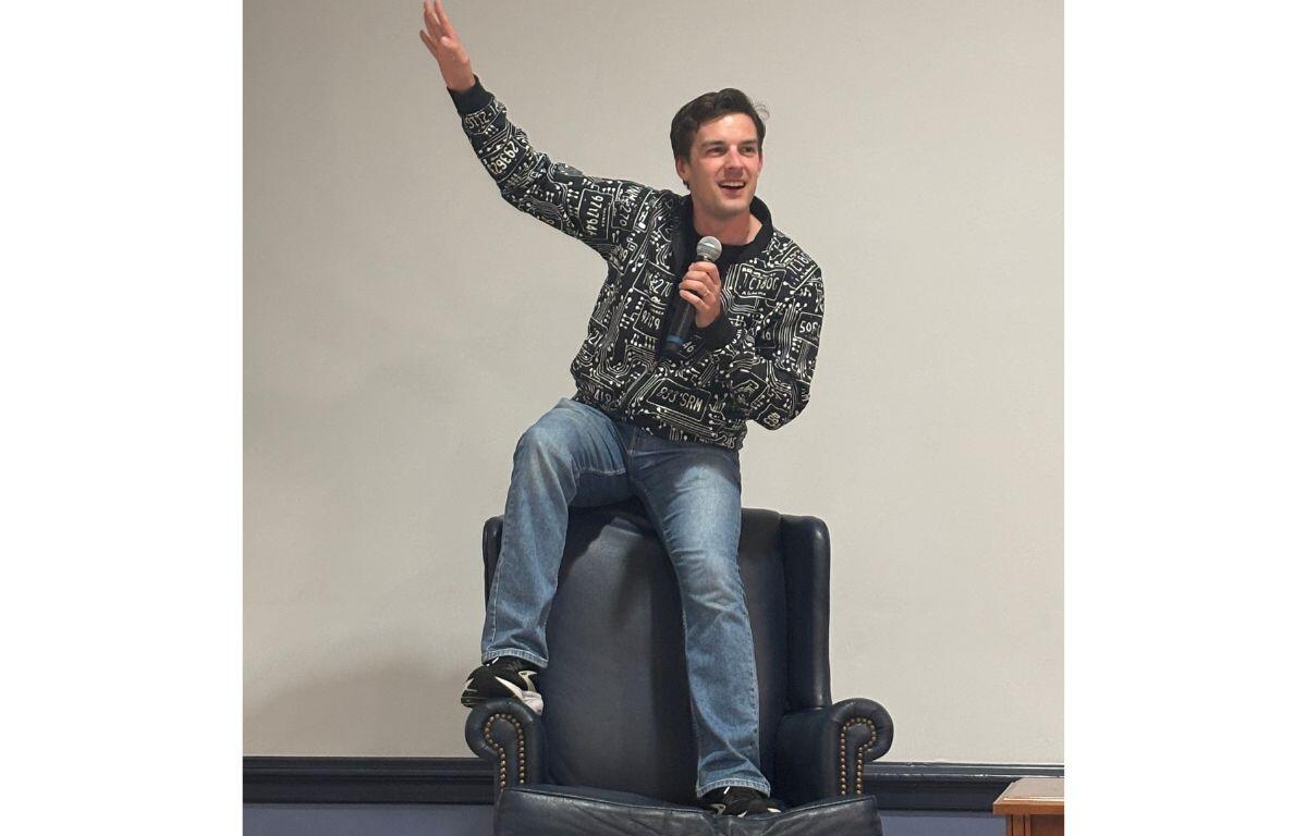 Man sits on a dark leather armchair, holding a microphone and raising one arm in a lively talk or performance.