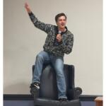 Man sits on a dark leather armchair, holding a microphone and raising one arm in a lively talk or performance.
