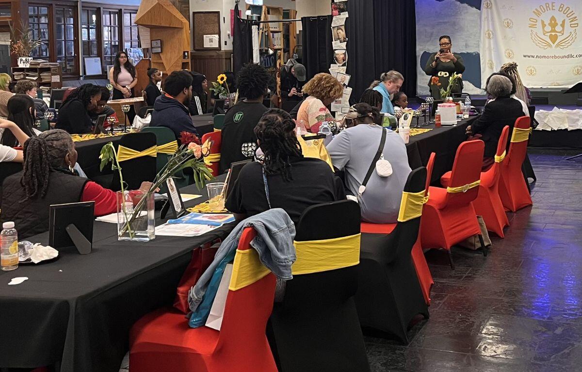 Group of people seated at long banquet tables in a hall, listening to a speaker on a stage with a banner reading 'No More Bound' in the background, flowers on tables and red chairs with yellow sashes.