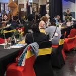 Group of people seated at long banquet tables in a hall, listening to a speaker on a stage with a banner reading 'No More Bound' in the background, flowers on tables and red chairs with yellow sashes.